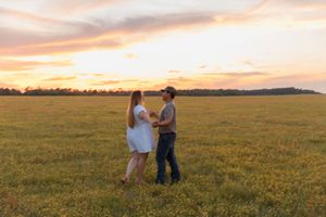 Couple holding hands in a golden field at sunset with colorful sky and tree line in the background.
