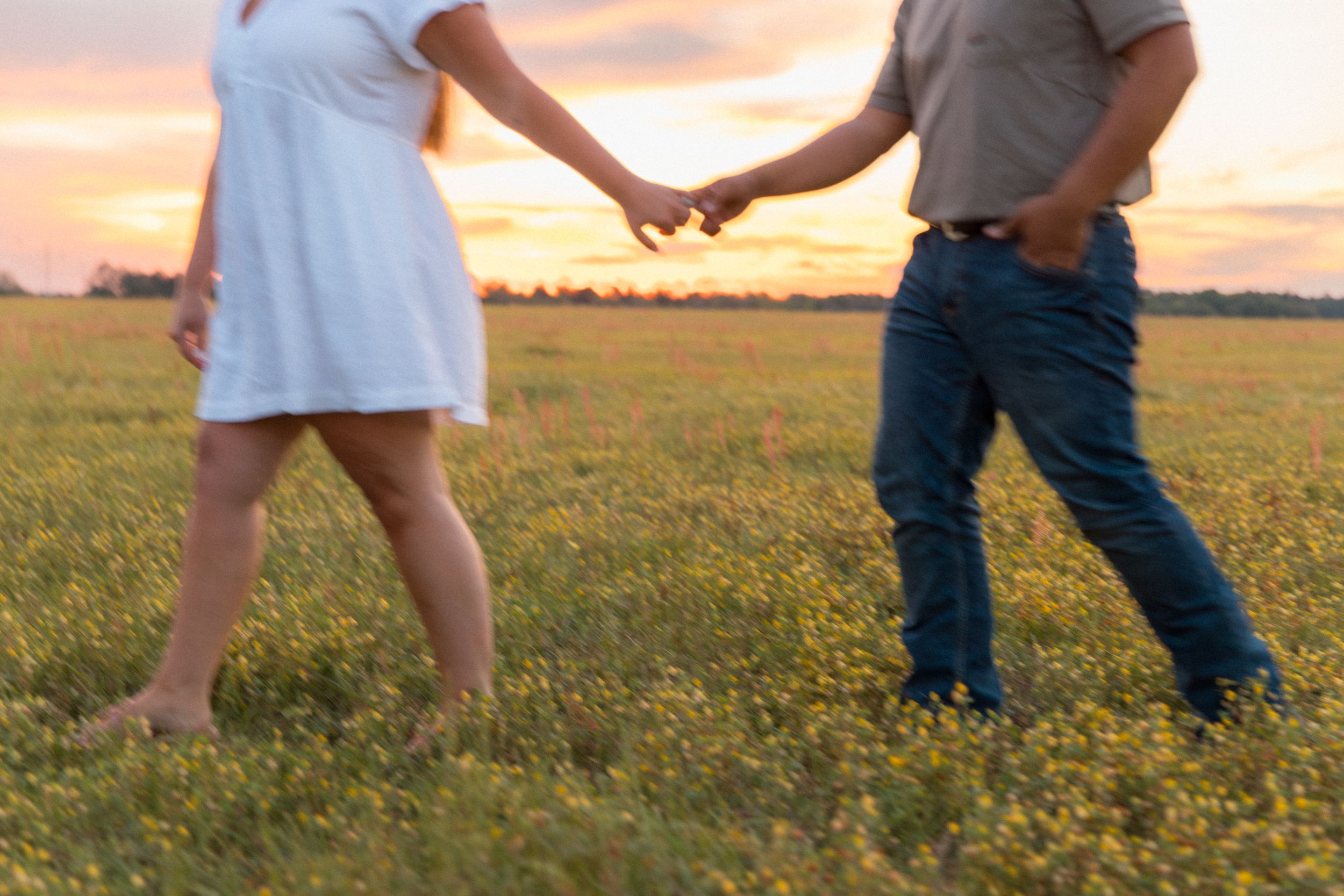 Couple holding hands walking through a golden flower field at sunset, woman in white dress, man in jeans.