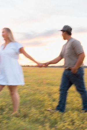 Couple holding hands walking through a green field at sunset, woman in white dress, man in jeans and cap.
