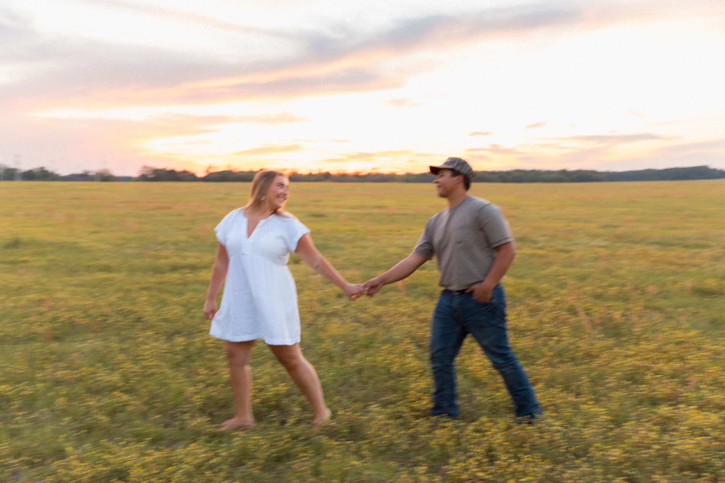 Couple holding hands walking through green field at golden hour sunset, woman in white dress, man in jeans.