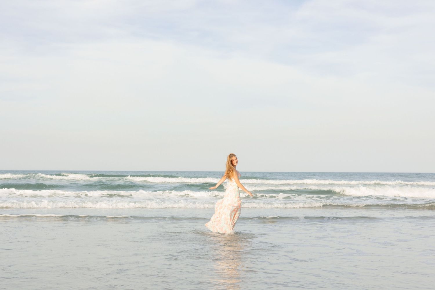 Young woman in floral maxi dress walking on sandy beach with ocean waves in background.