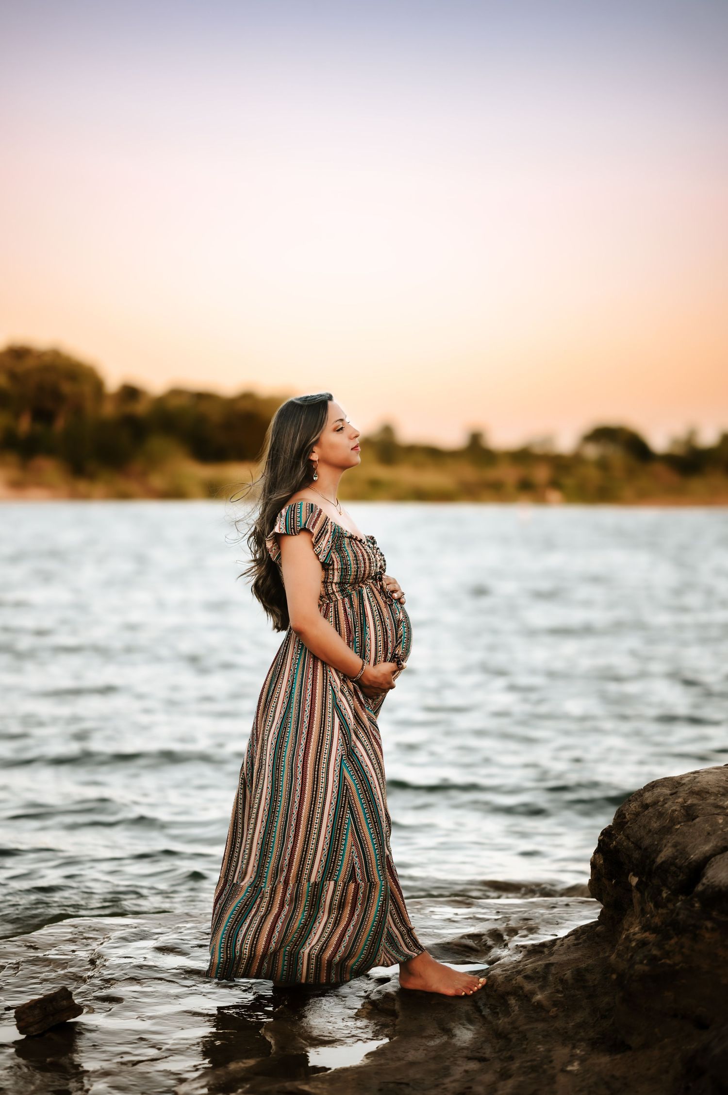 Pregnant woman in colorful striped maxi dress standing barefoot by lake gazing up at sunset sky.