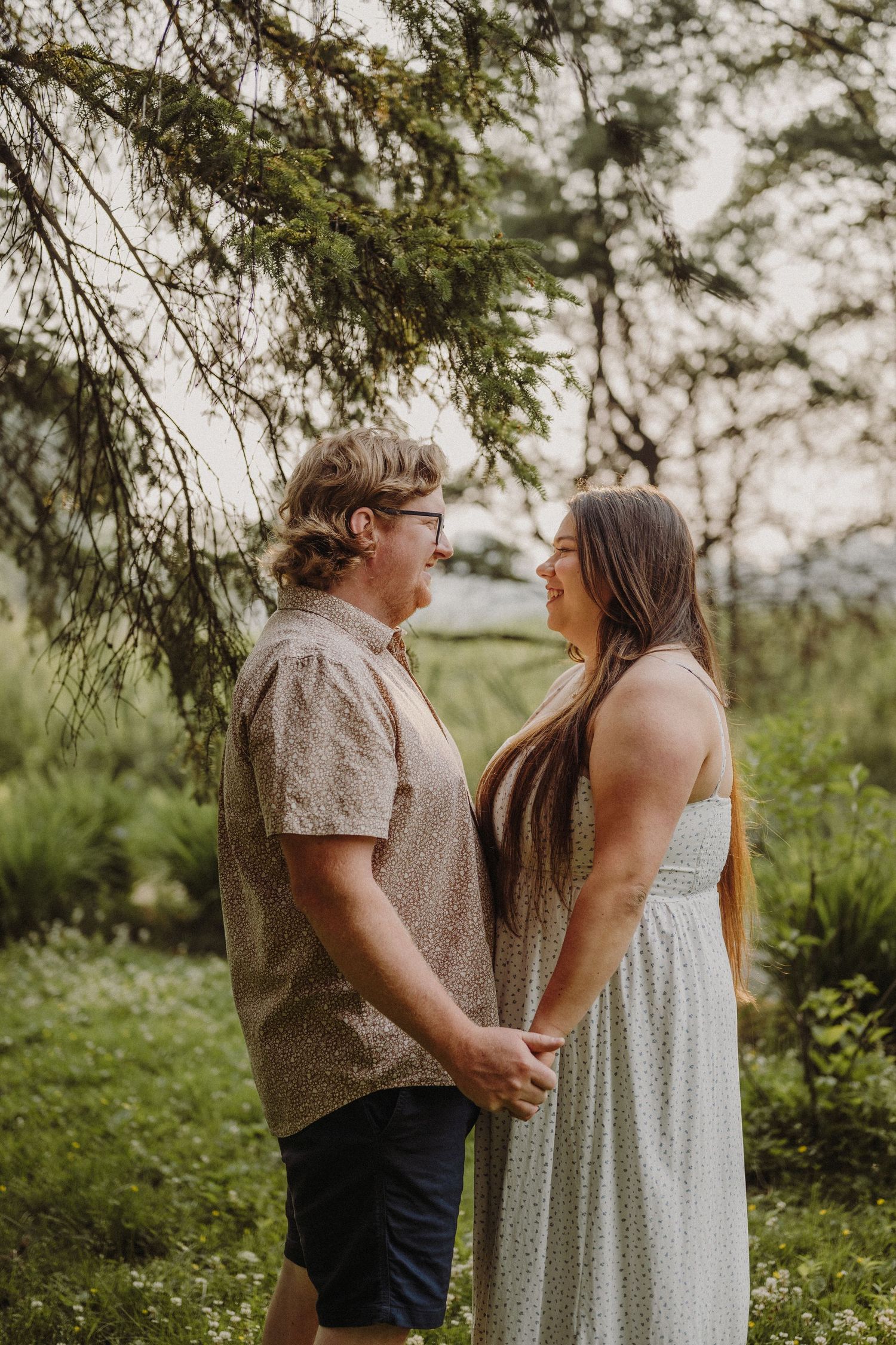 Couple holds hands in lush green field with power tower and dramatic cloudy sky at golden hour.