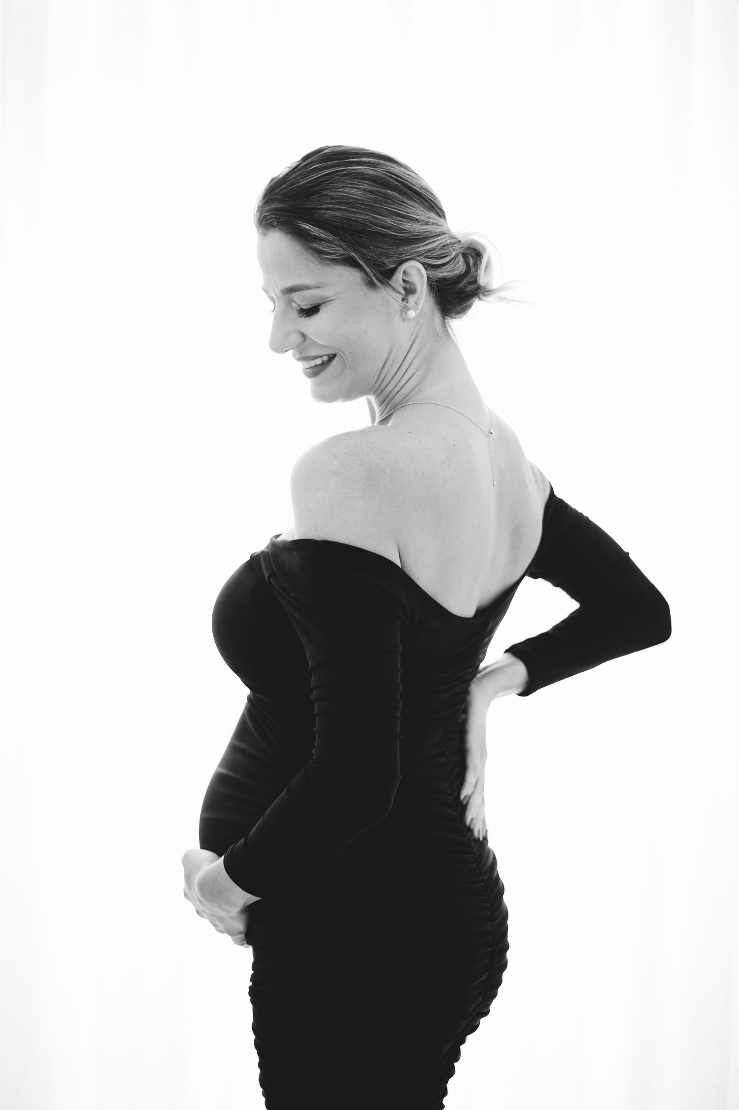 Black and white maternity photo of a smiling woman in an elegant off-shoulder black dress cradling her baby bump.