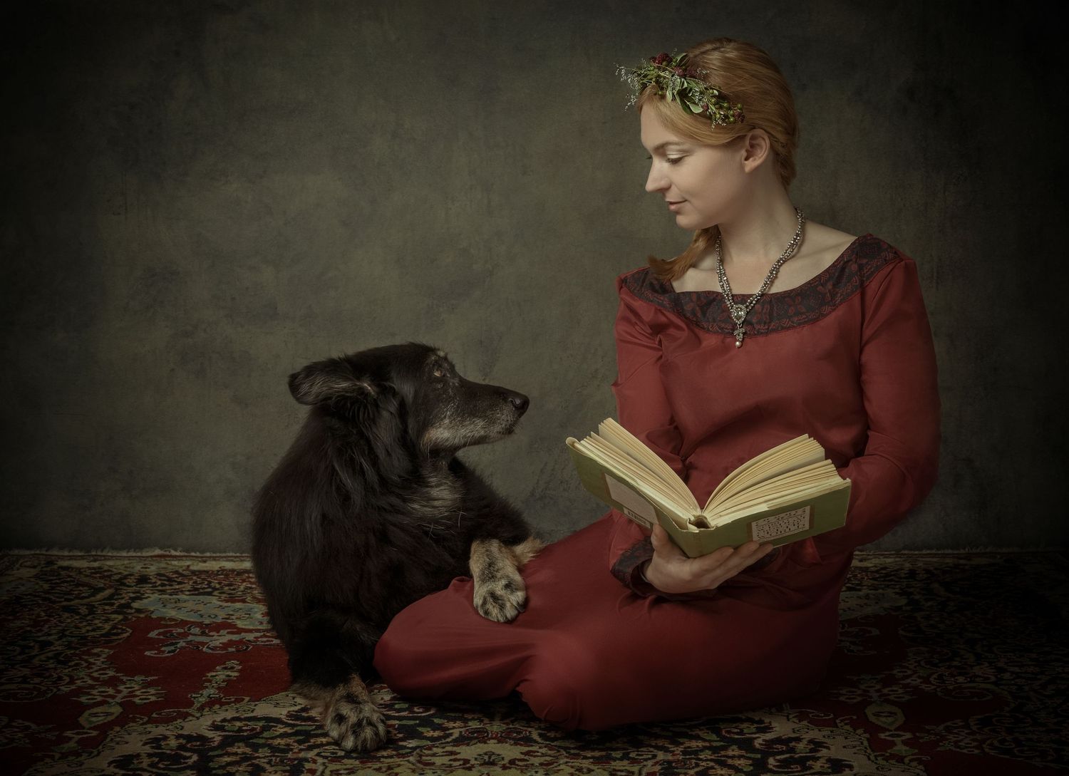 A young woman in red medieval dress reads a book to her attentive black dog while sitting on a patterned rug.