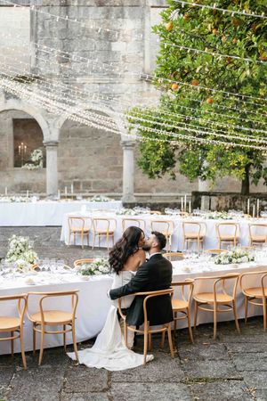 Long wedding reception table setup outdoors with wooden chairs and white linens under stone archways.