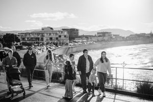 People walking along a seaside promenade in black and white, with ocean waves and coastal buildings in the background.