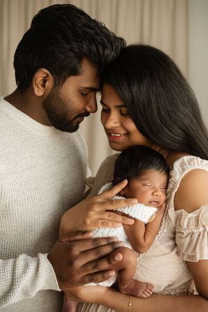 A young South Asian couple tenderly holds their sleeping newborn baby, smiling warmly in a soft, neutral-toned studio setting.