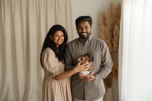 Happy couple smiling and holding their newborn baby in a bright, neutral-toned photography studio setting.