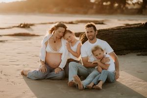 Pregnant woman with family sitting on beach at golden hour sunset, smiling couple with two children in casual white and denim.