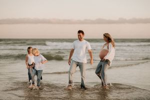 Pregnant woman and family walking on beach at sunset, children playing in ocean waves.