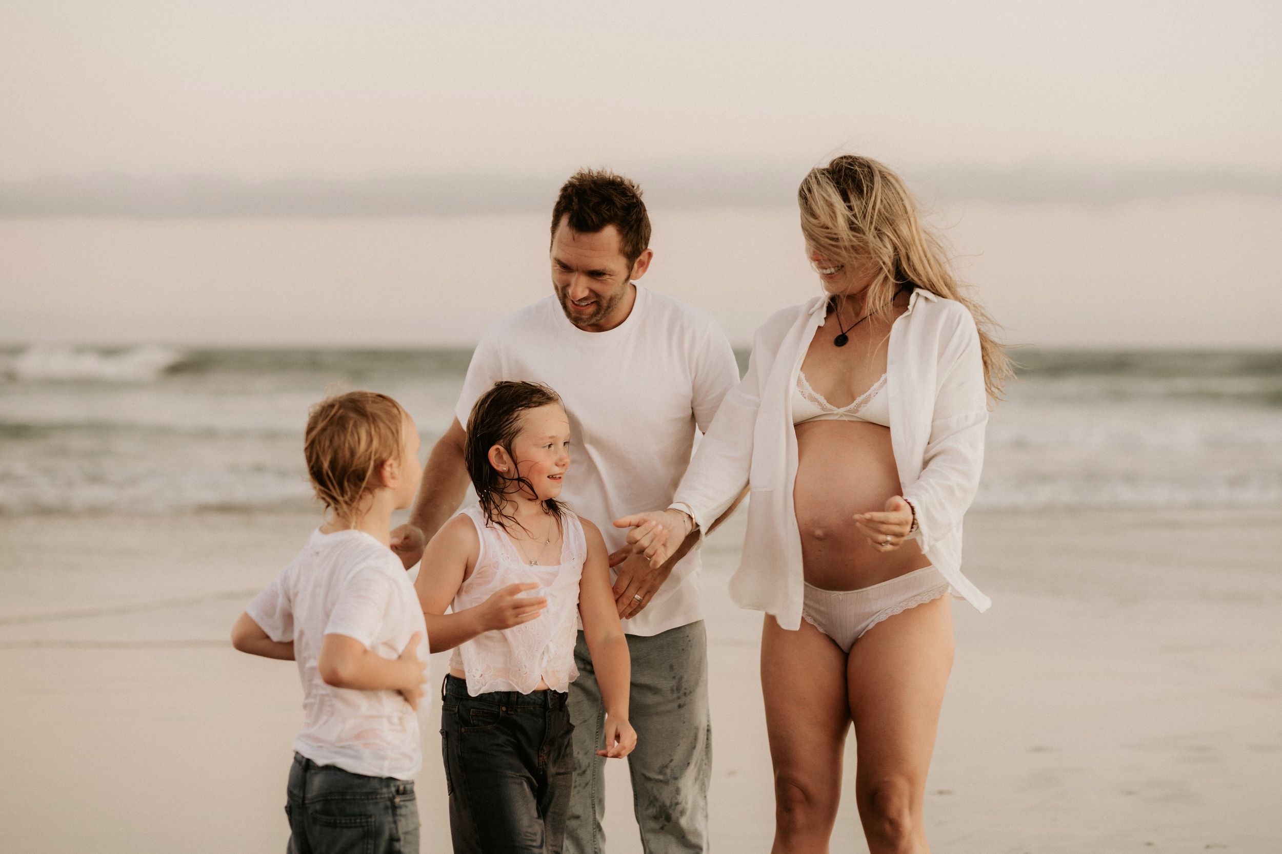 Pregnant woman with family on beach, husband and two children holding hands by ocean waves at sunset.