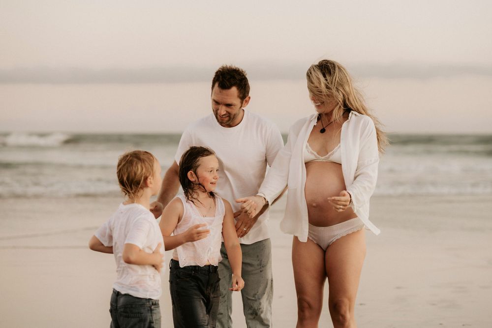 Pregnant woman with family on beach, husband and two children holding hands by ocean waves at sunset.