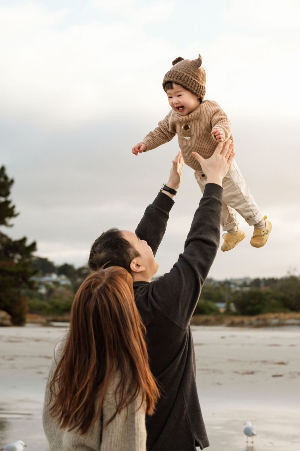 Parents joyfully tossing smiling baby in brown knit outfit at a beach, creating a heartwarming family moment.