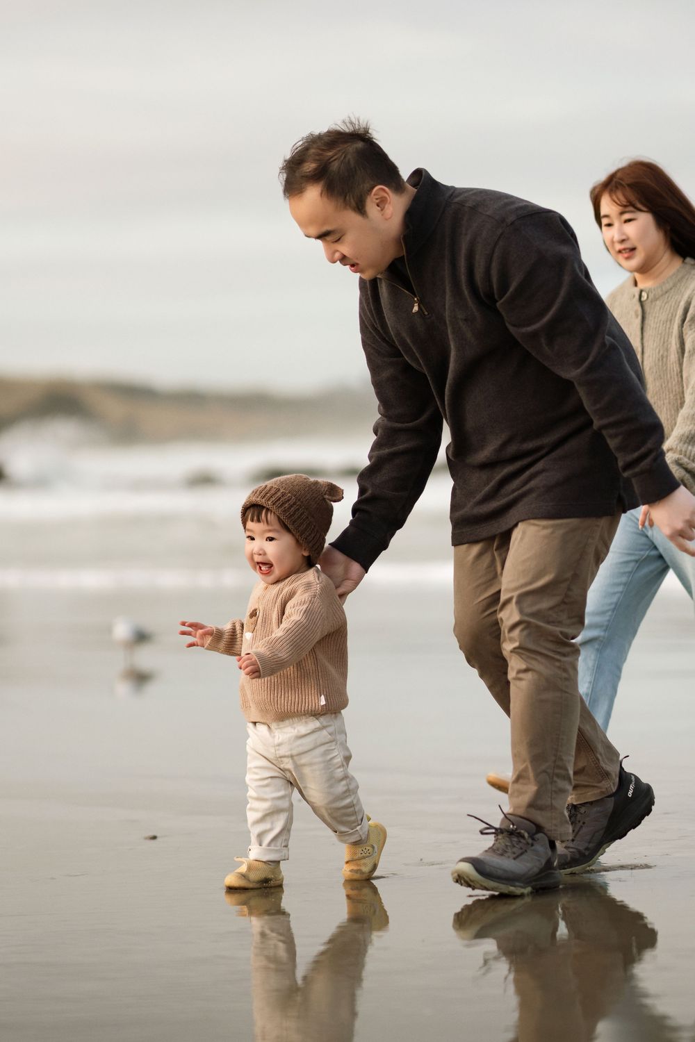 Family with toddler walking on a reflective beach, parents holding child's hands on a cloudy day.