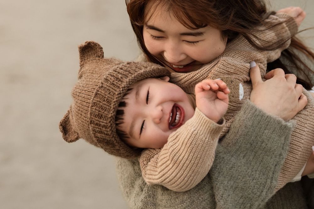 A joyful mother hugging her laughing toddler wearing a bear ear knit hat and sweater outdoors.