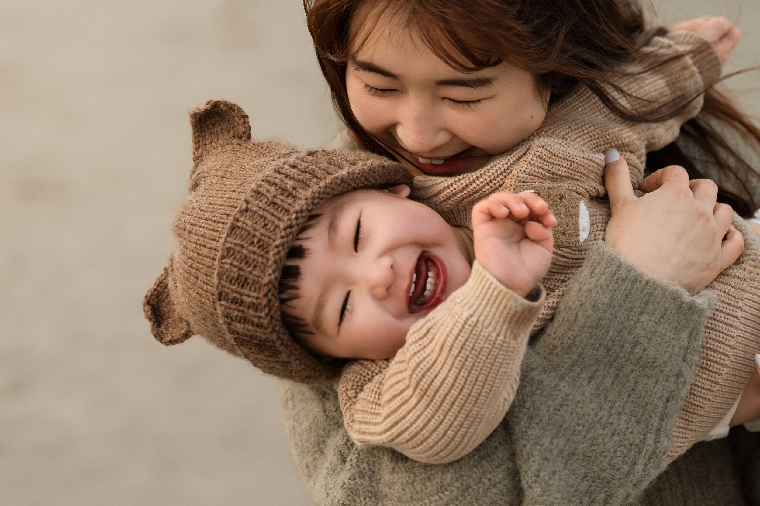 A joyful mother hugging her laughing toddler wearing a bear ear knit hat and sweater outdoors.