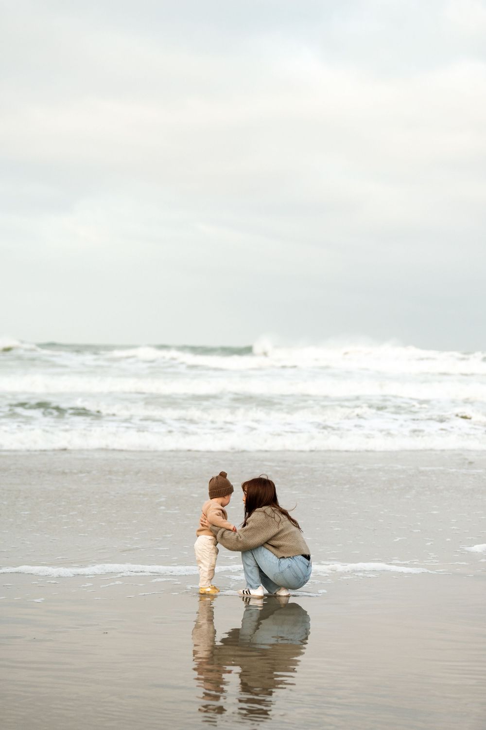 A mother crouches on a wet beach facing her toddler near ocean waves on a cloudy day.