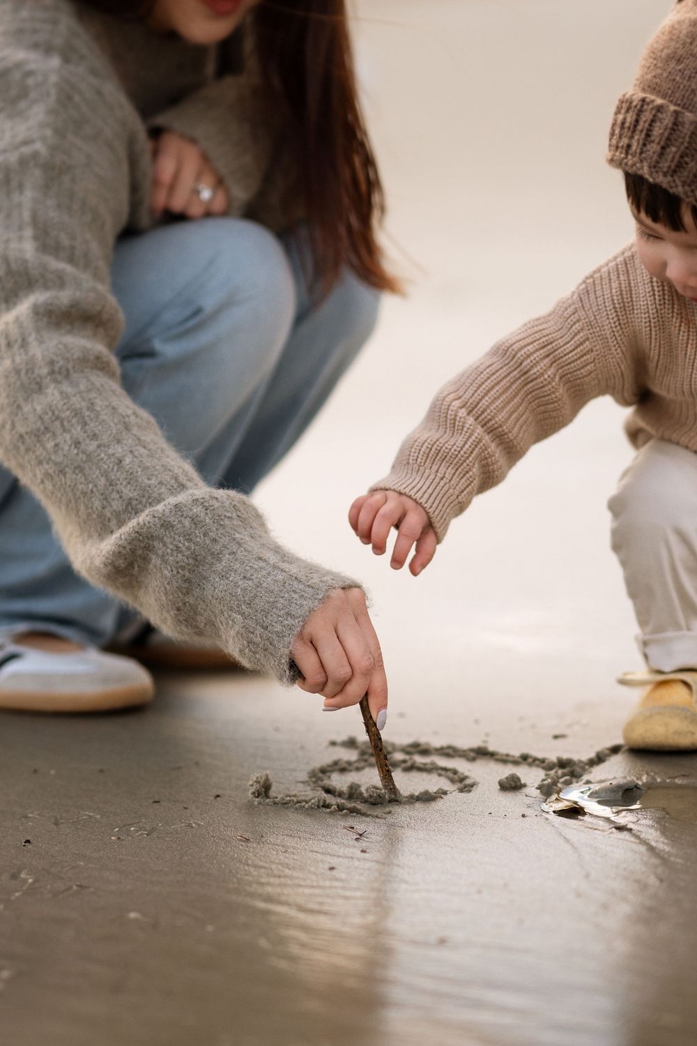 Mother and child drawing a heart in wet sand at the beach using a stick, both wearing cozy sweaters.