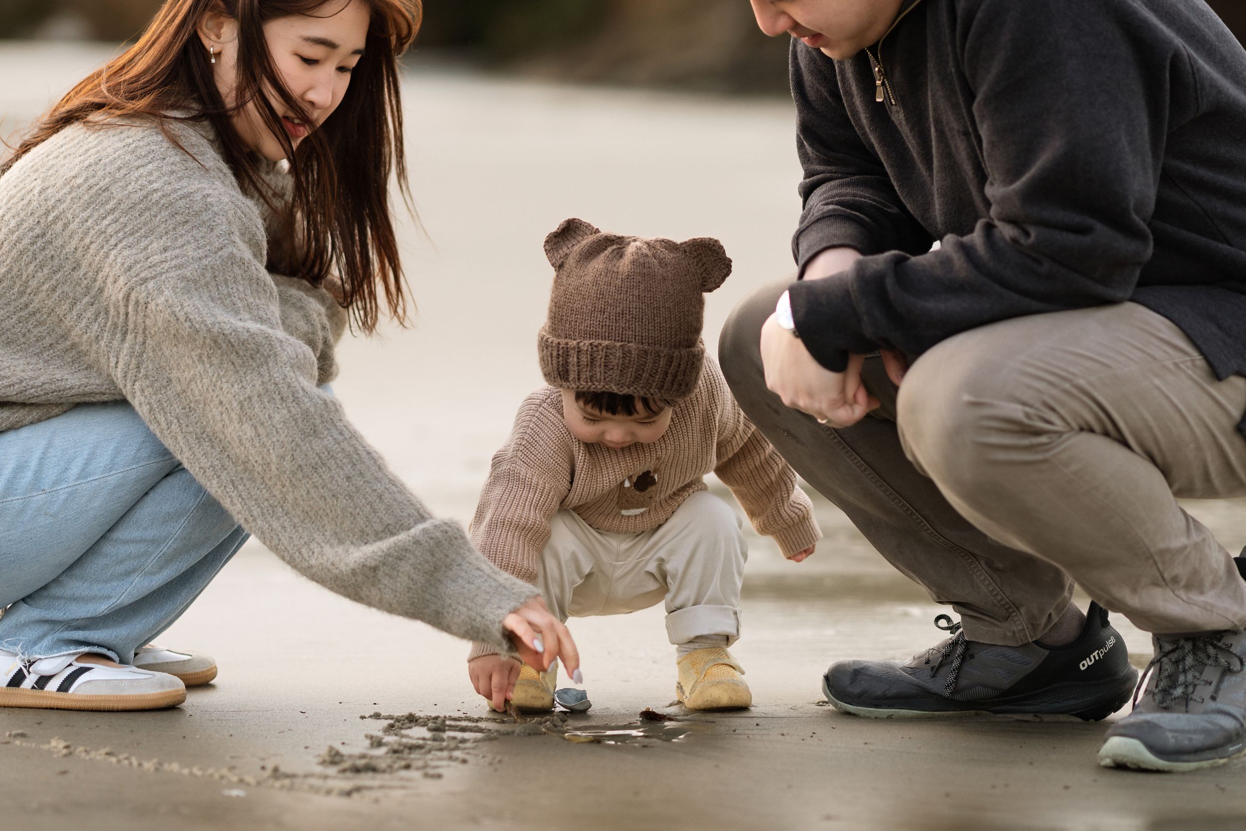 Young family with toddler crouching outdoors, parents helping child explore ground together.