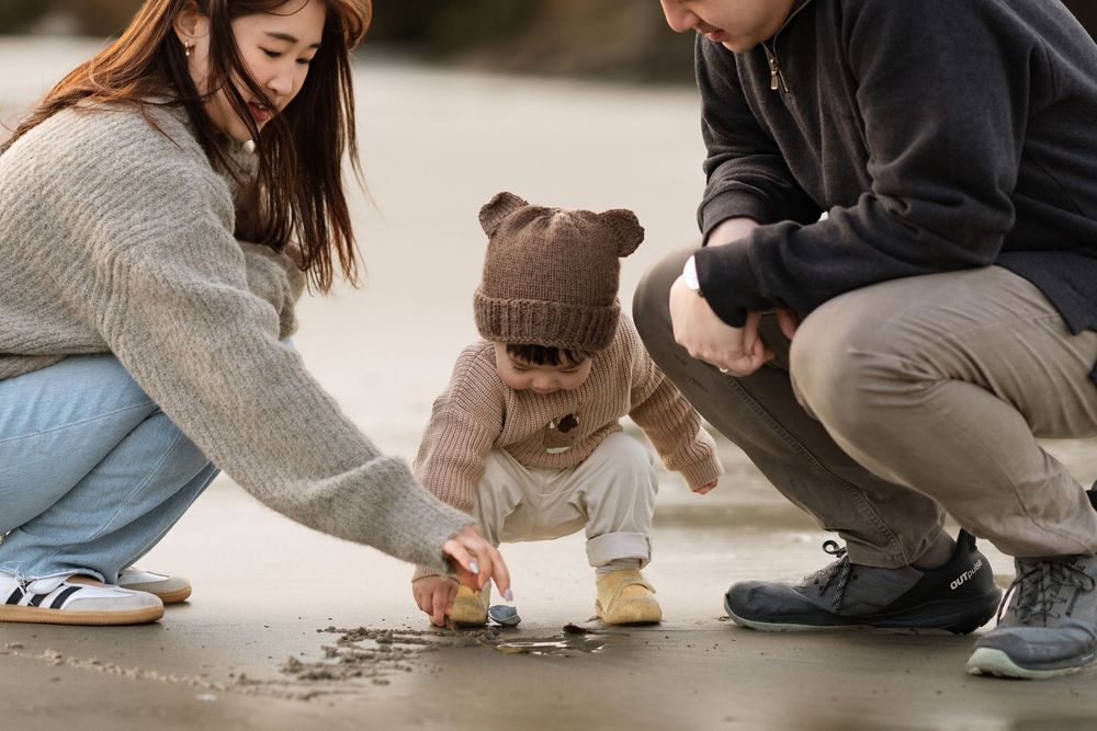 Young family with toddler crouching outdoors, parents helping child explore ground together.