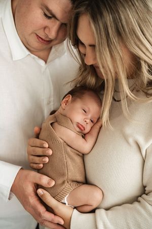 Two parents tenderly holding their sleeping newborn baby dressed in a cozy brown knit outfit.