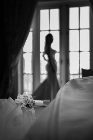 Black and white photo of a bride silhouetted in a window with a bouquet in the foreground.