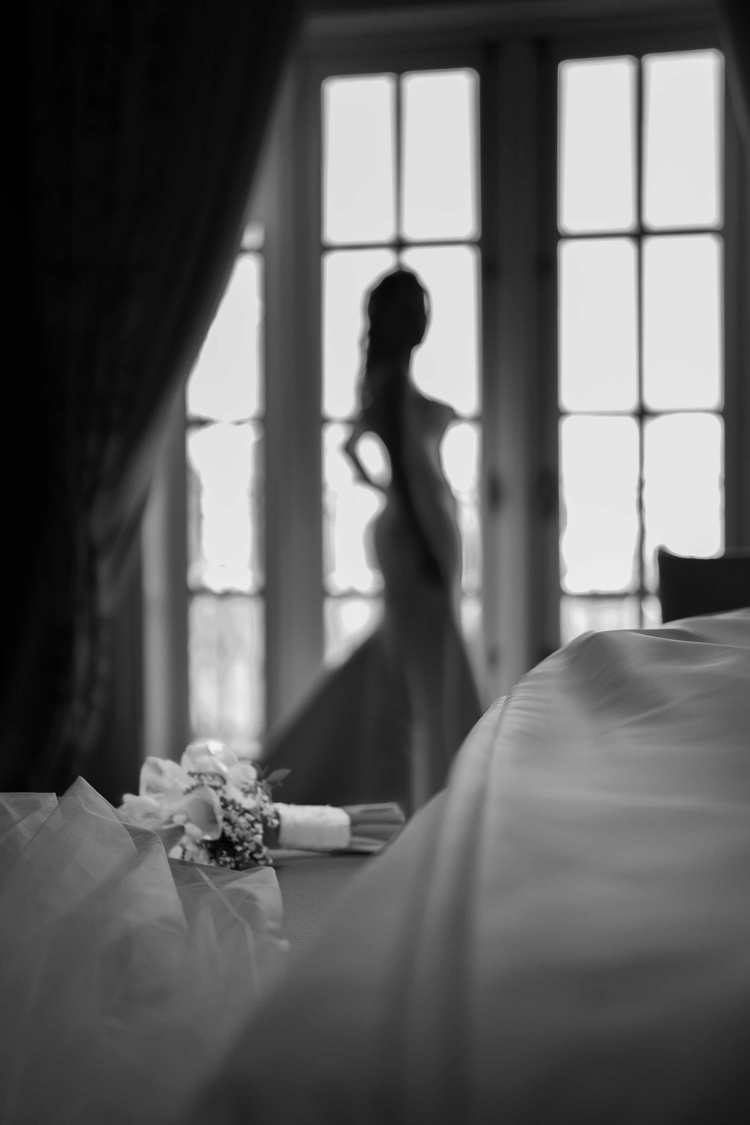 Black and white photo of a bride silhouetted in a window with a bouquet in the foreground.