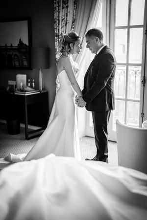 Black and white photo of bride and groom holding hands by window in elegant room, wedding dress train flowing.