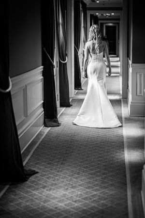 Bride in elegant mermaid wedding gown walks down a grand hotel corridor in a black and white photo.