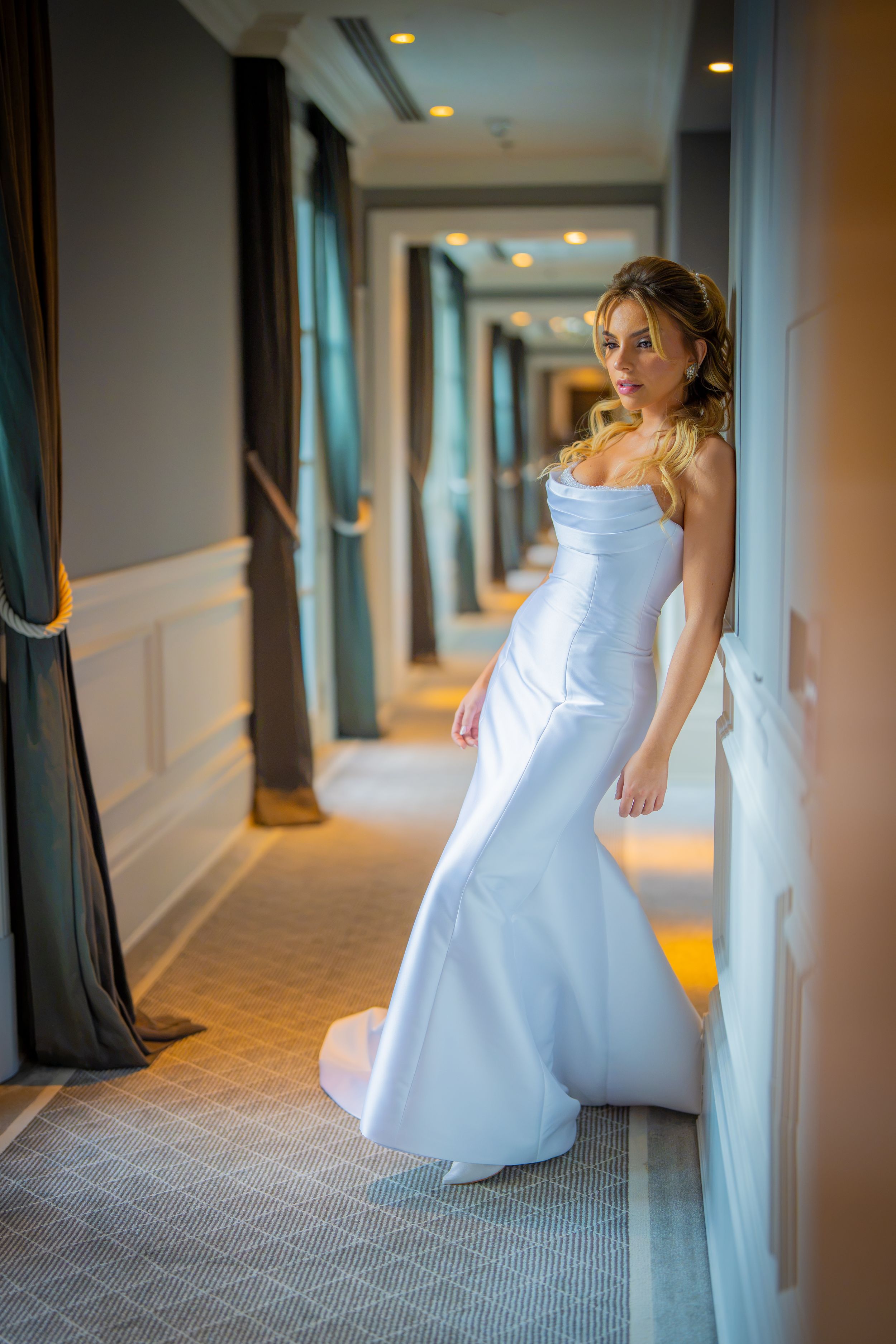 Elegant bride in a white satin mermaid gown posing in a luxurious hotel corridor with teal curtains.