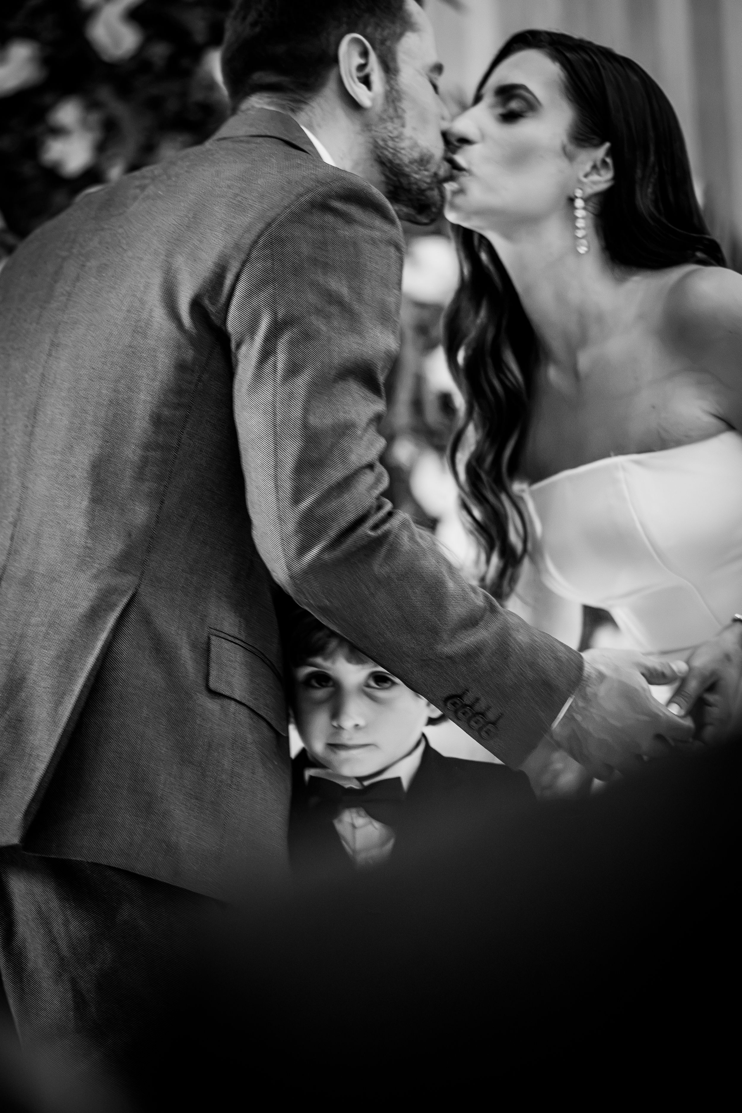 Black and white wedding photo of couple kissing while young boy peeks between them during ceremony.