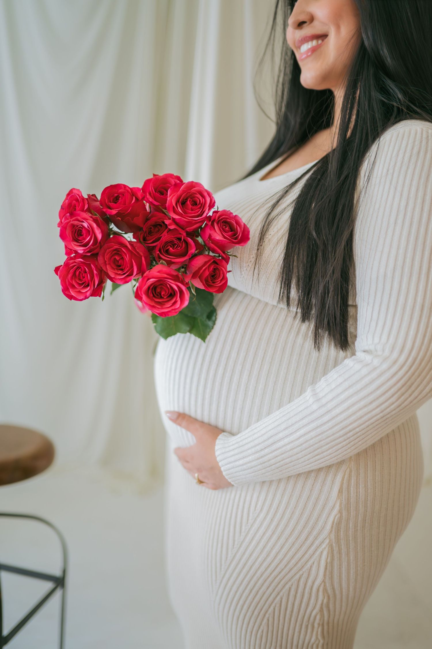 Pregnant woman in white ribbed dress smiles while holding bouquet of red roses indoors Asheboro NC