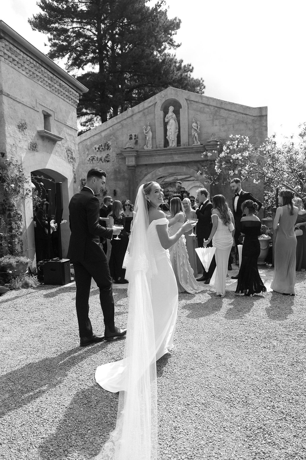 Black and white photo of a bride in a flowing veil and groom in tuxedo at an elegant outdoor wedding venue.