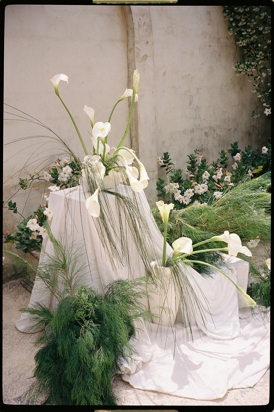 White calla lilies and green ferns arranged on draped white fabric pedestals against a stone wall.