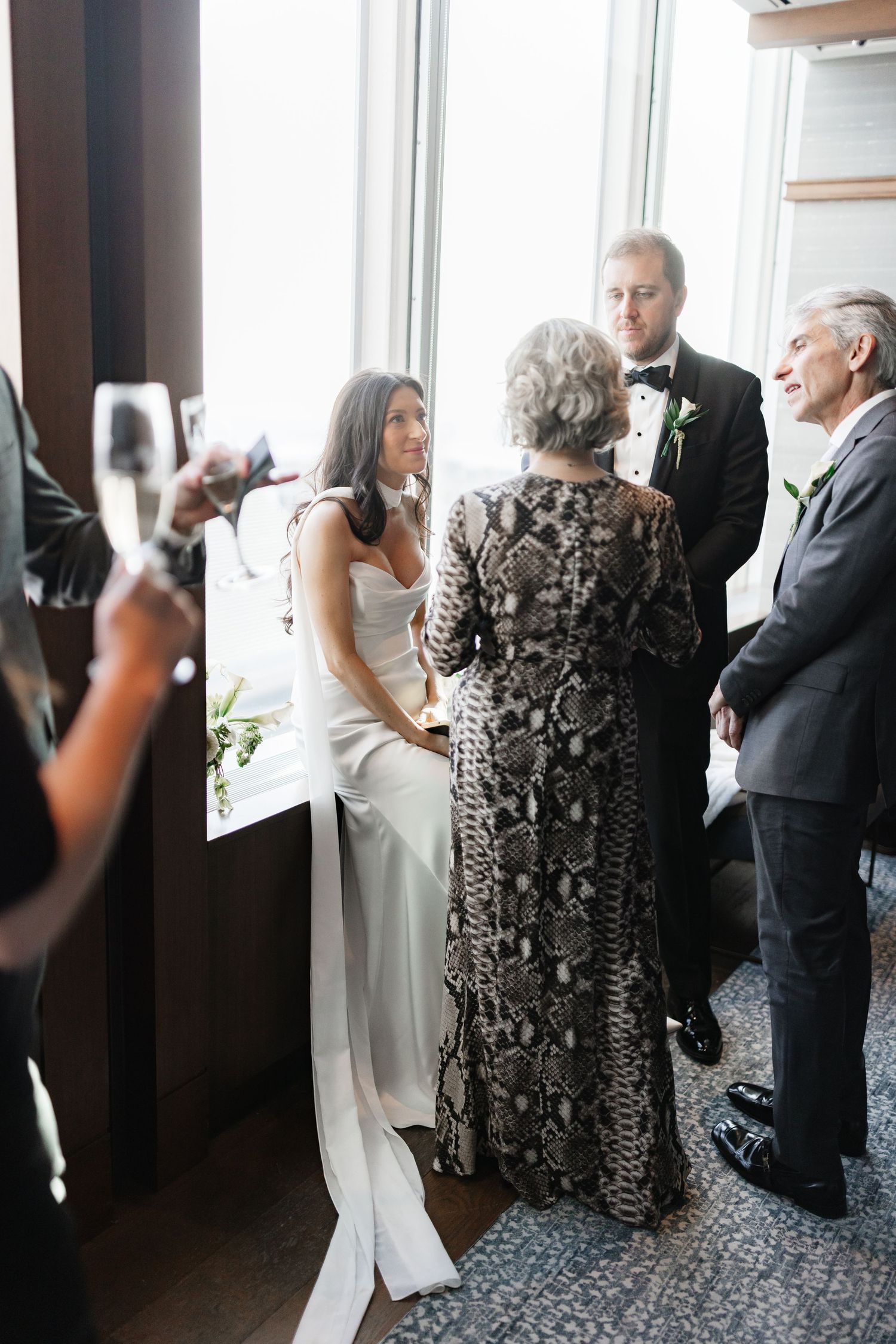 Bride in white gown chats with guests in elegant attire at an indoor wedding reception near large windows.