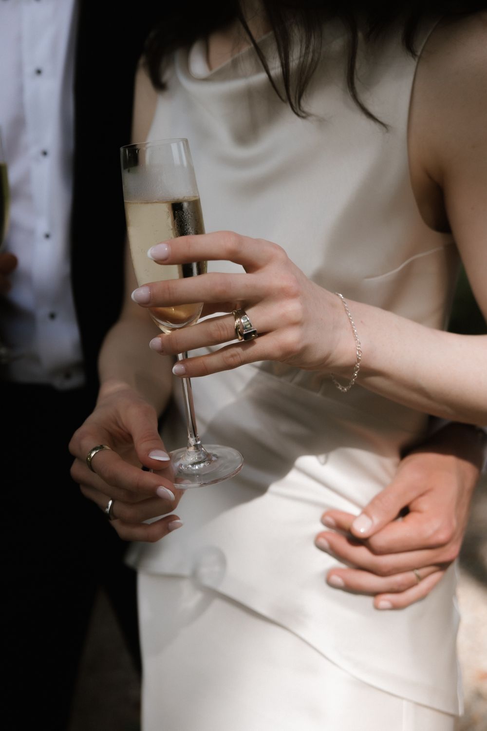 Bride in white satin gown holding champagne flute, adorned with rings and bracelet, groom's hand resting on her waist.