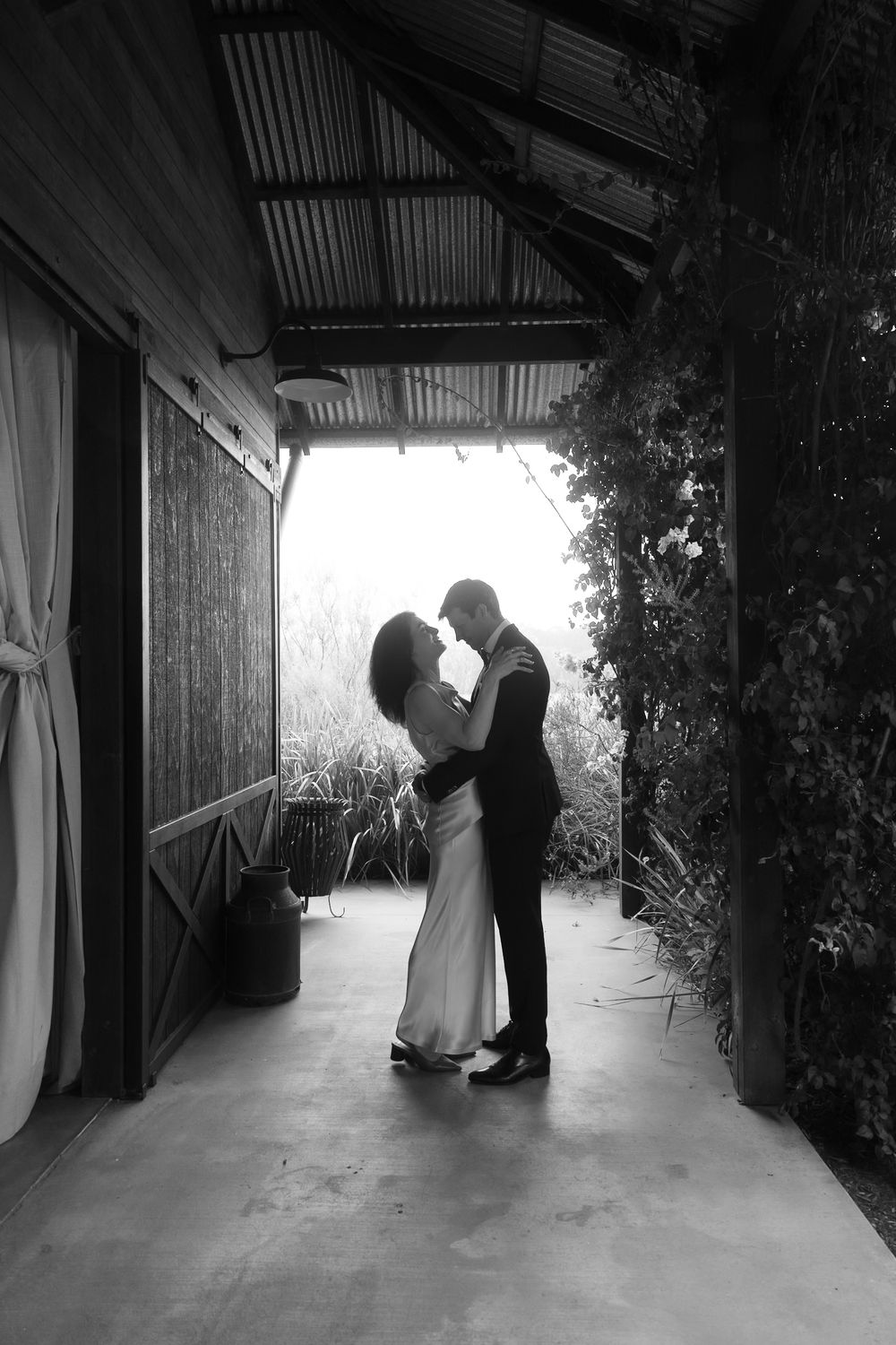 Black and white photo of a couple sharing a romantic embrace in a rustic covered walkway surrounded by lush greenery.