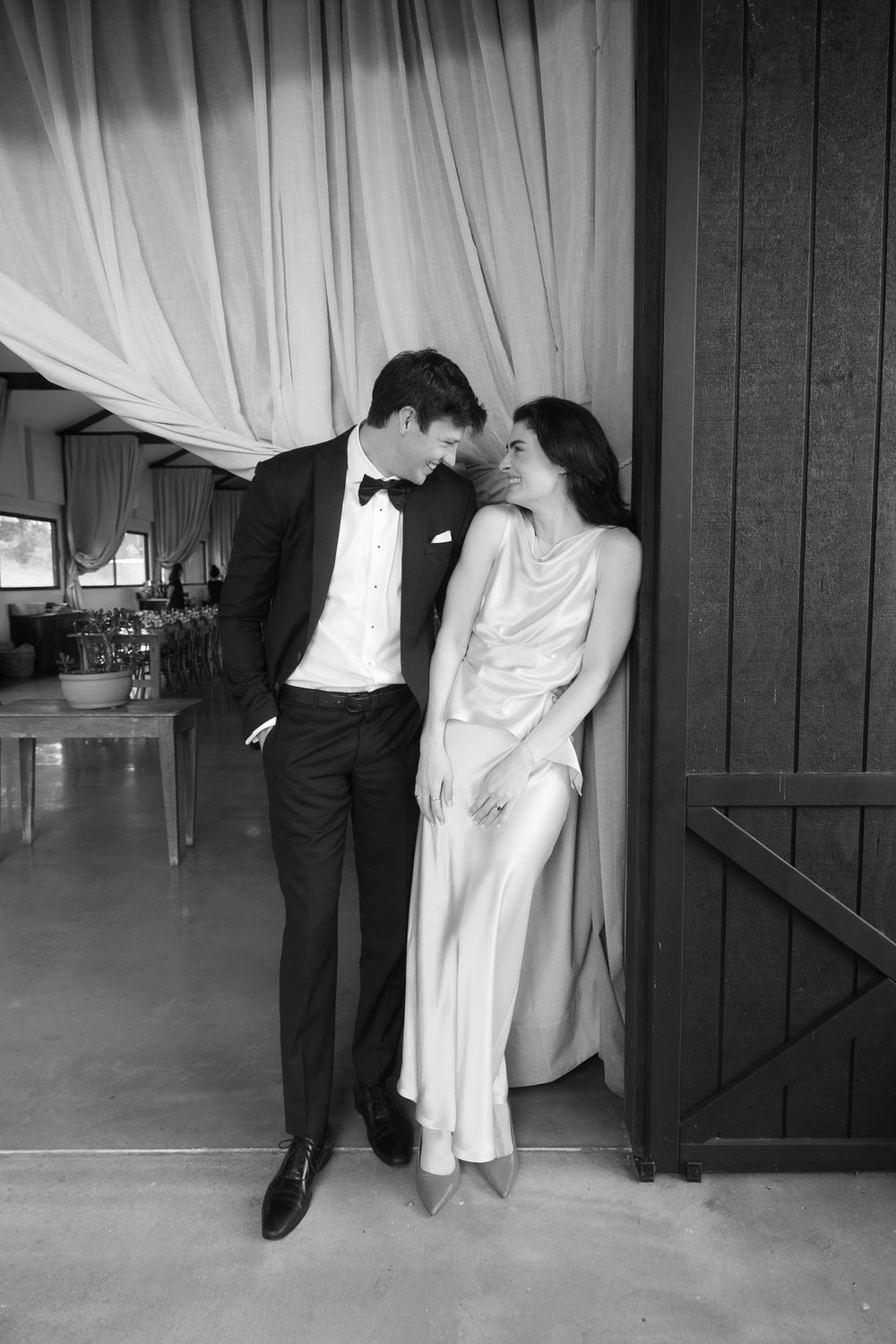 Black and white photo of an elegant couple in formal attire sharing an intimate moment by draped curtains.