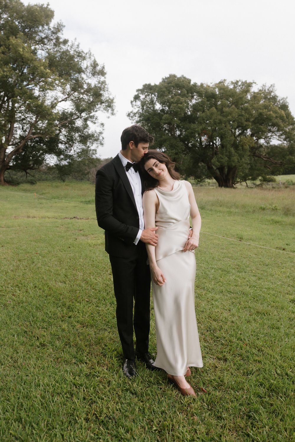 Couple in wedding attire embracing on green grass field, groom in black tuxedo, bride in elegant ivory satin gown.