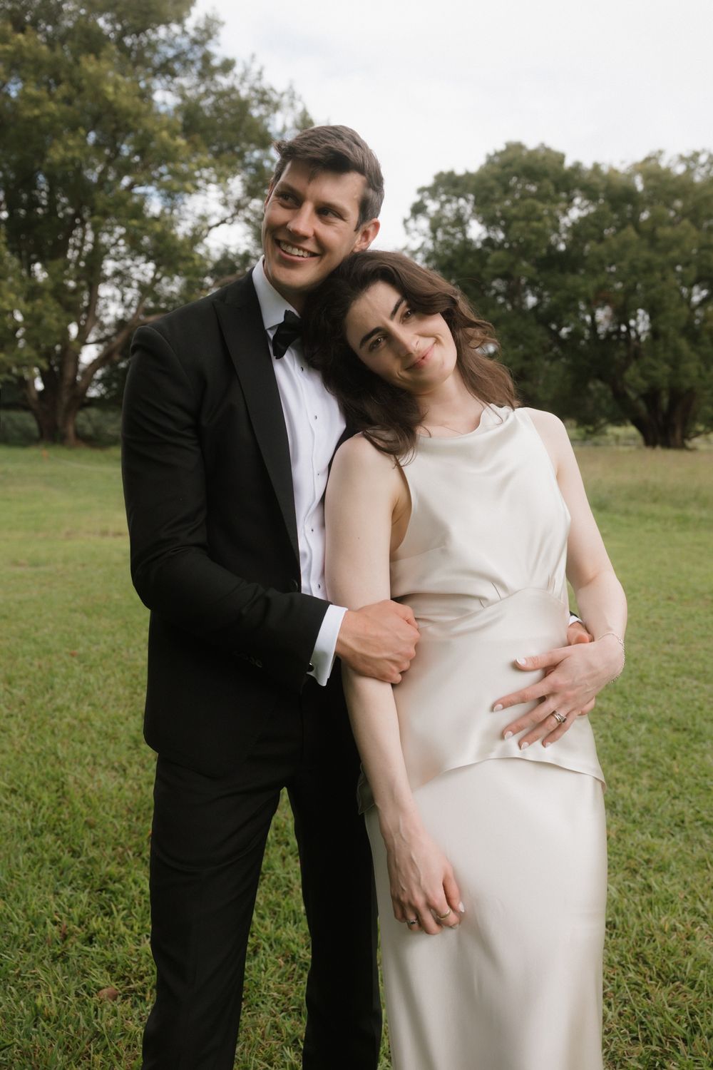 A smiling couple in wedding attire embrace outdoors, man in black tuxedo, woman in white satin gown, green trees behind.