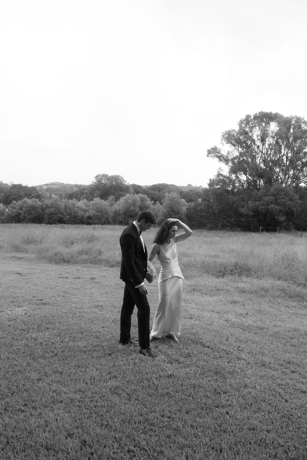 Black and white photo of a bride and groom holding hands in a grassy field with trees in the background.