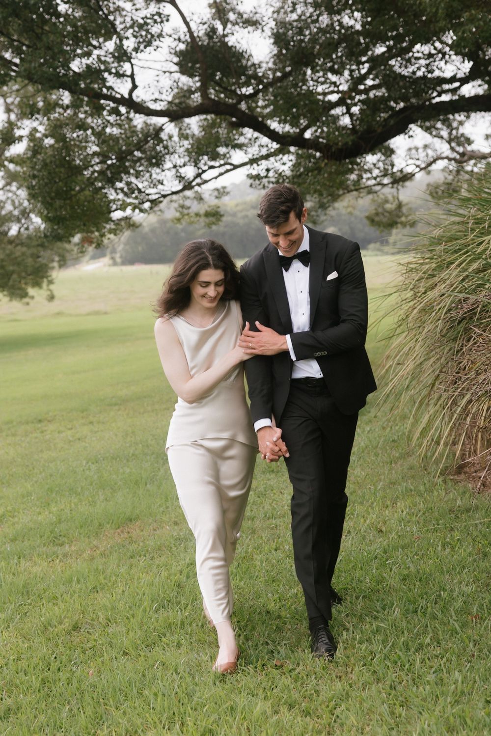 Elegant couple walking hand in hand on lush green grass, woman in white jumpsuit and man in black tuxedo with bow tie.