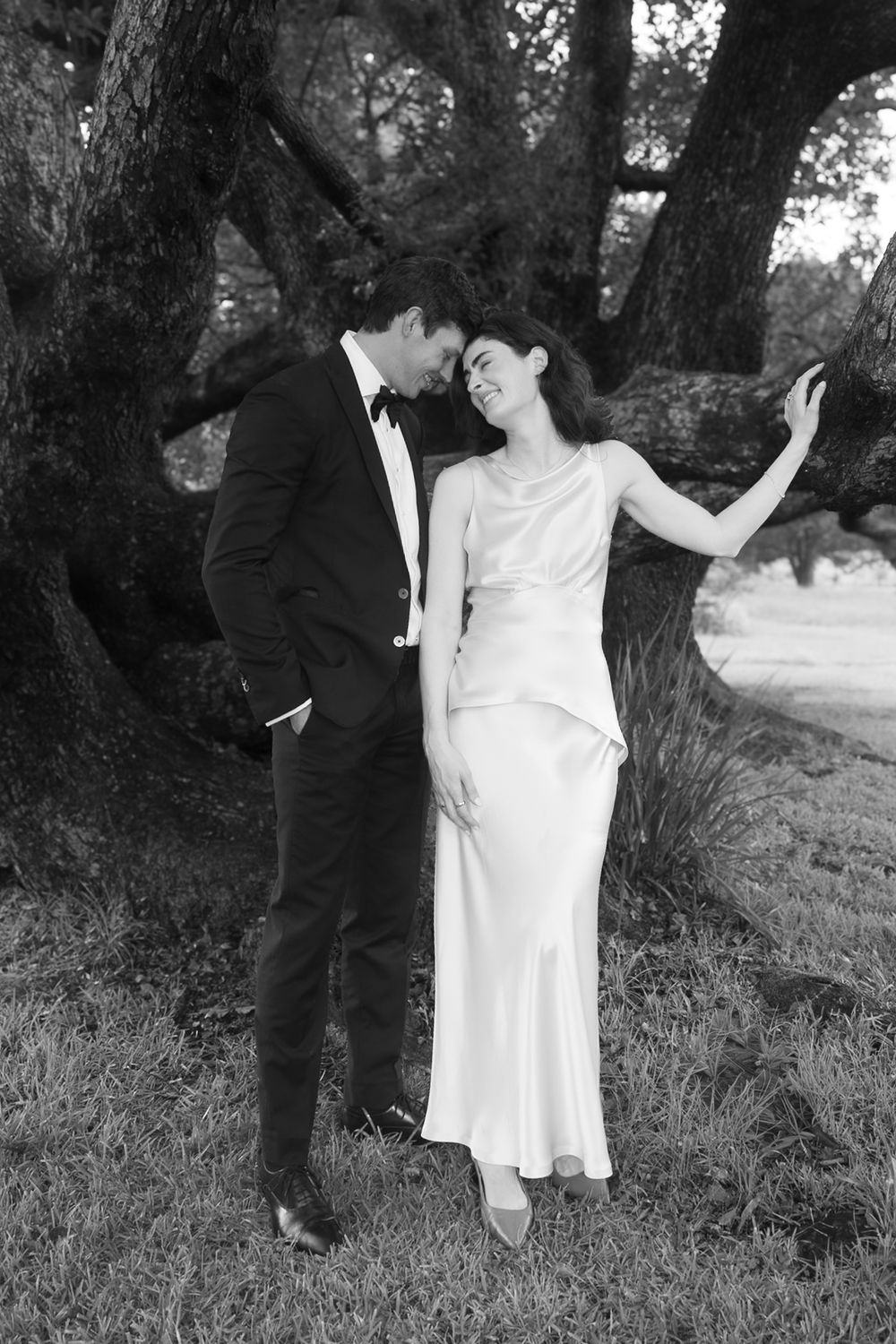 Black and white photo of an elegant couple in formal wear embracing near a large tree outdoors.
