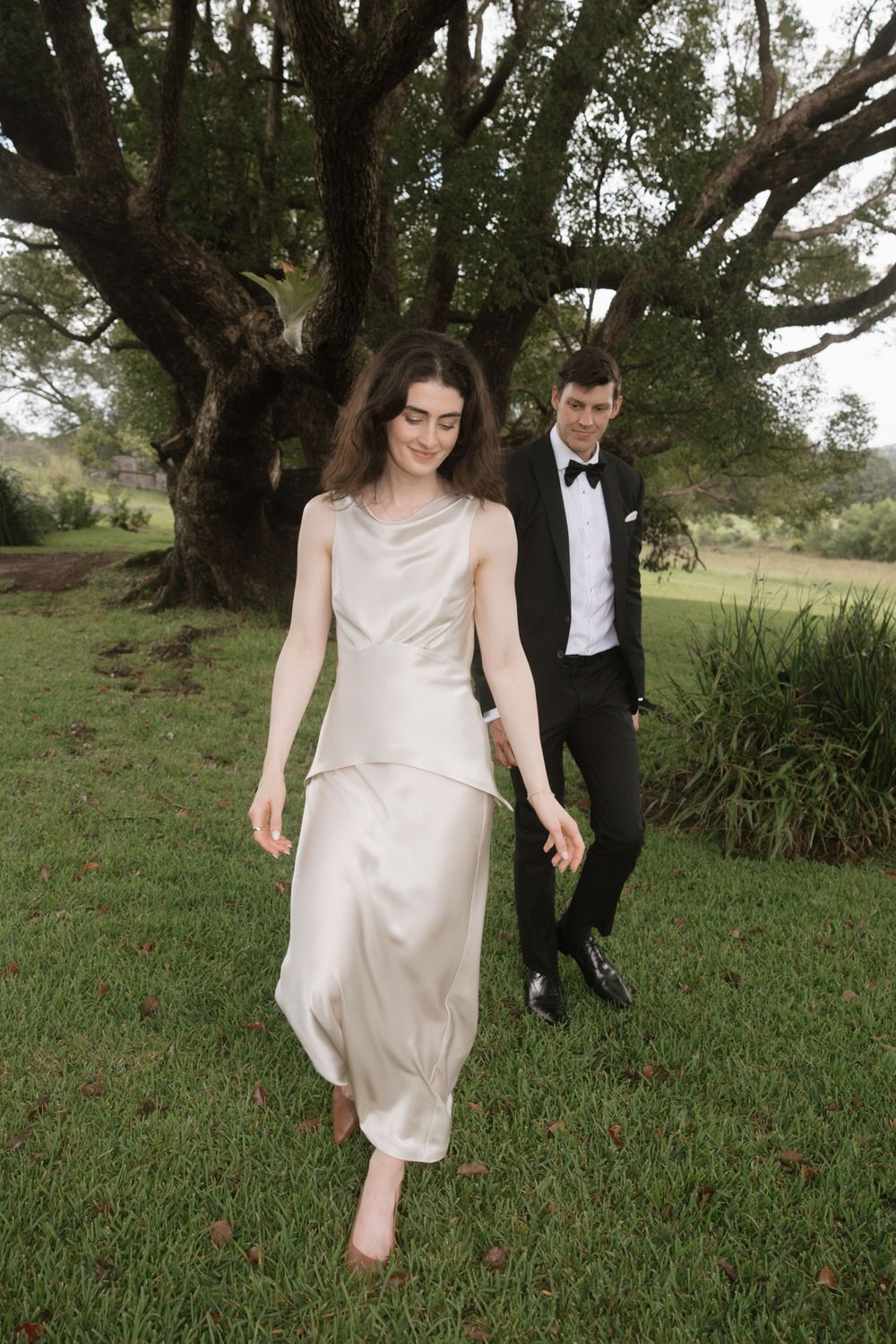 Bride in flowing white satin wedding dress walks ahead of groom in black tuxedo on lush green lawn.