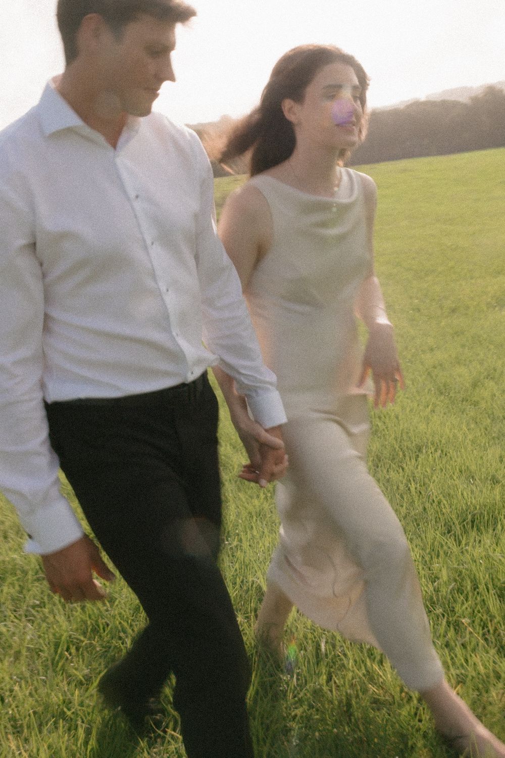 A couple in elegant attire walks hand-in-hand through a lush green field at sunset.