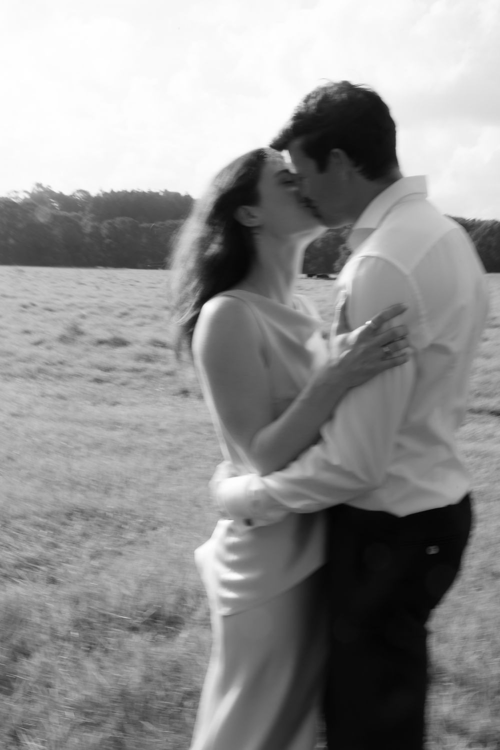 Black and white photo of a romantic couple kissing in a grassy field, woman in white dress embracing man in white shirt.