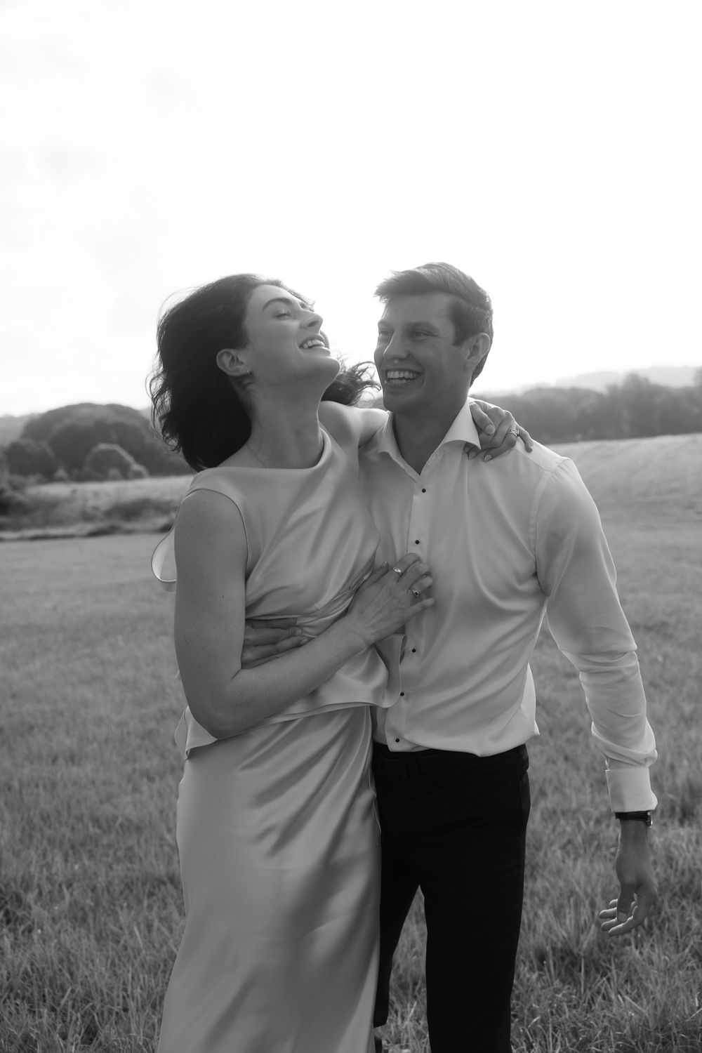 Black and white photo of a laughing couple embracing in a field, woman in elegant dress, man in white shirt.