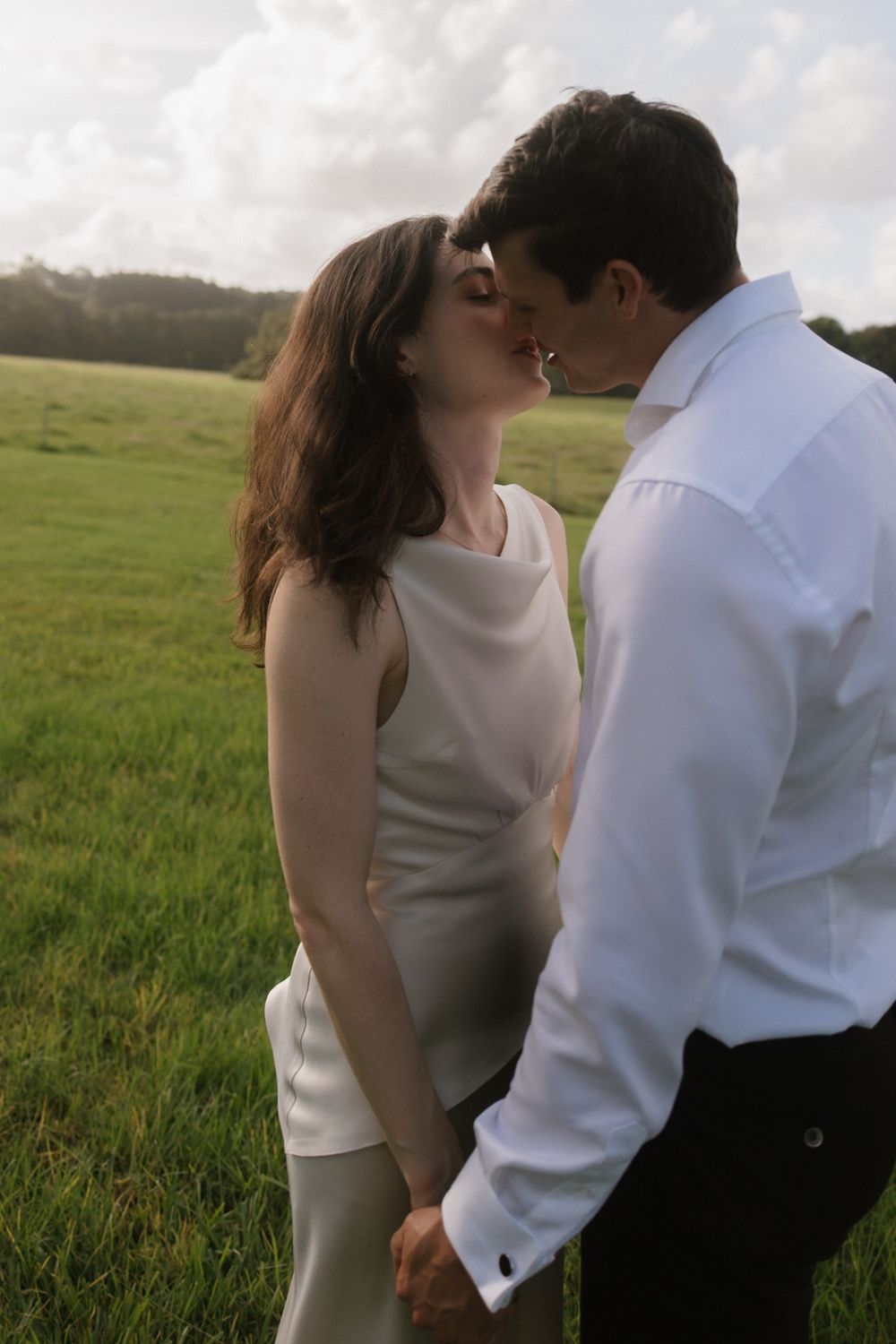 A couple shares a romantic kiss in a lush green field, dressed in elegant white attire on a cloudy day.