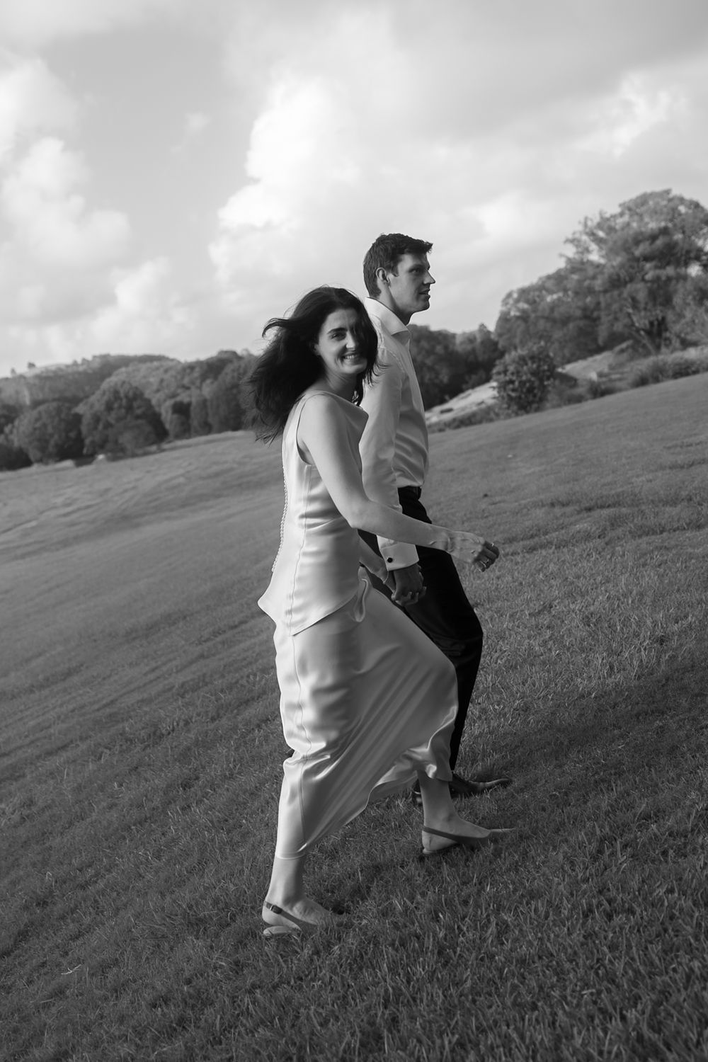 Black and white photo of a couple in wedding attire walking hand in hand across a lush green field.
